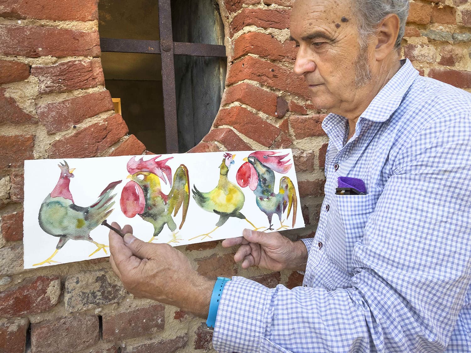 Fotografia dell'artista che presenta in mano l'acquerello di varie galline in fila. L'artista è serio e sullo sfondo è presente una finestra ovale circondata dai mattoni.